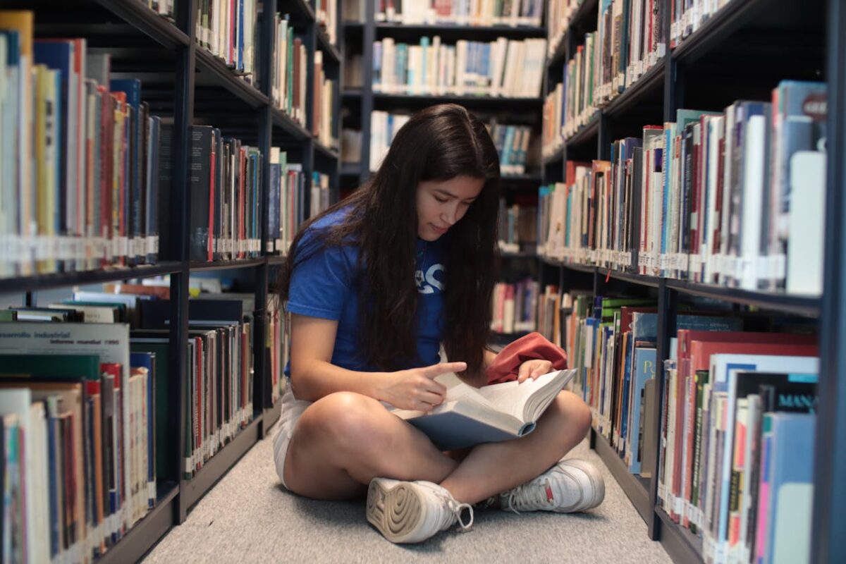 Woman sitting on floor reading a book in library aisle with shelves full of books.